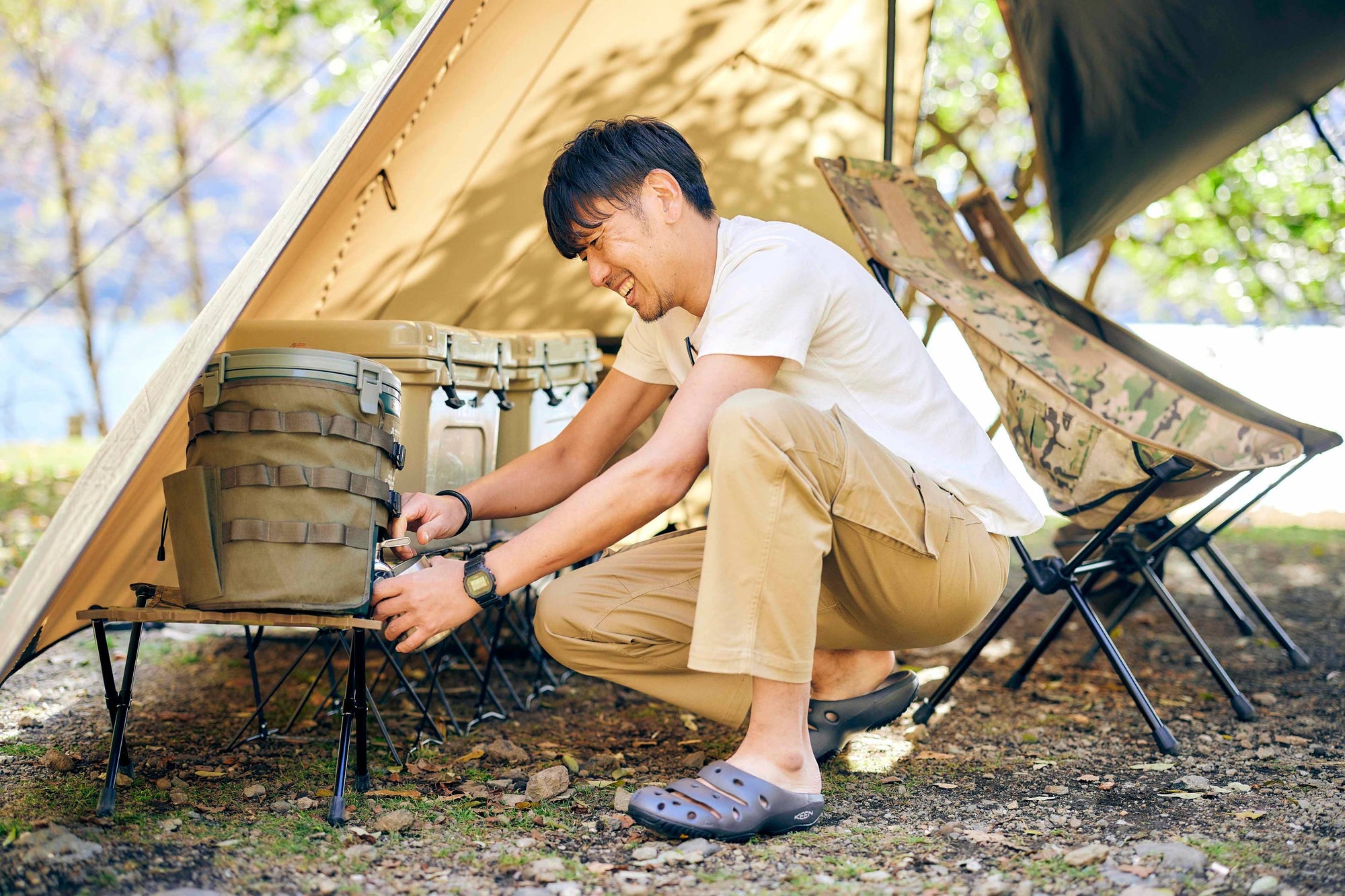 A man smiling, dispensing water from a small cooler inside a tent, wearing a pair of KEEN sandals.