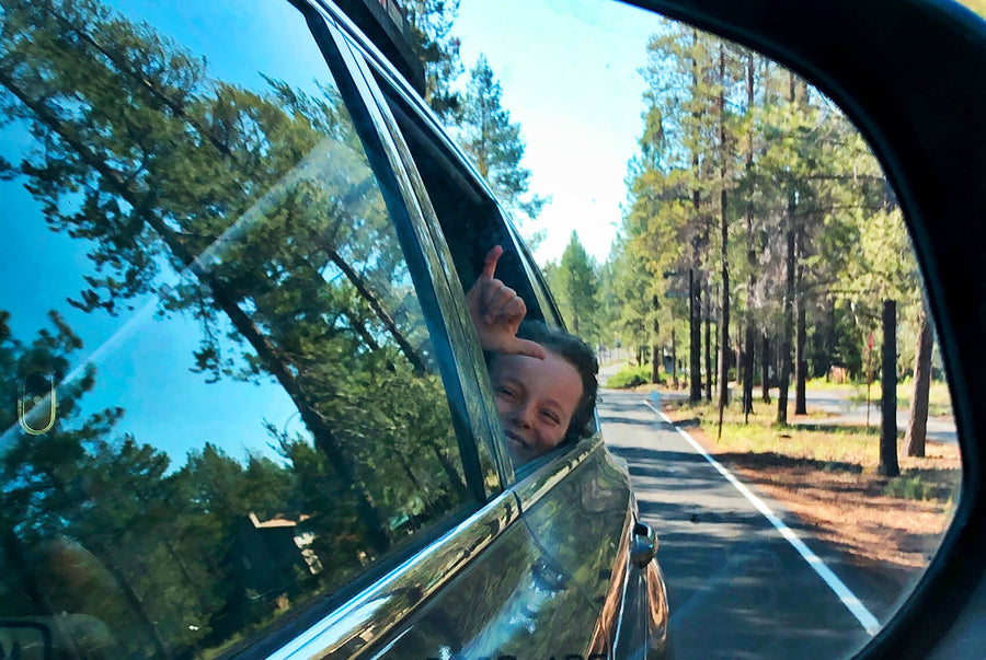 A kid poking their head out of a car window and smiling, with a forest scene behind them.