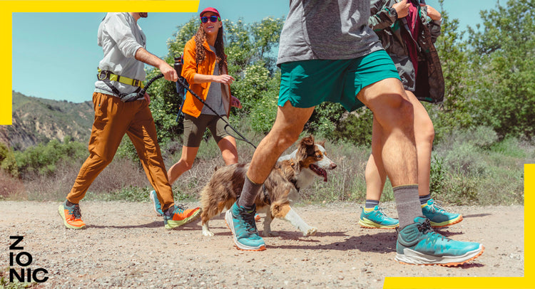 knee down view of 4 people and a dog hiking on flat dirt road wearing Zionics