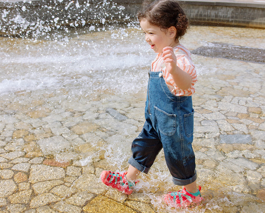 Toddler girl in denim overalls wearing Hyperport sandals and walking through large fountain play area. 