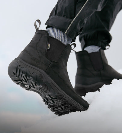 A knee down shot of a person wearing black slip-on insulated boots hiking through a blanket of thick snow.