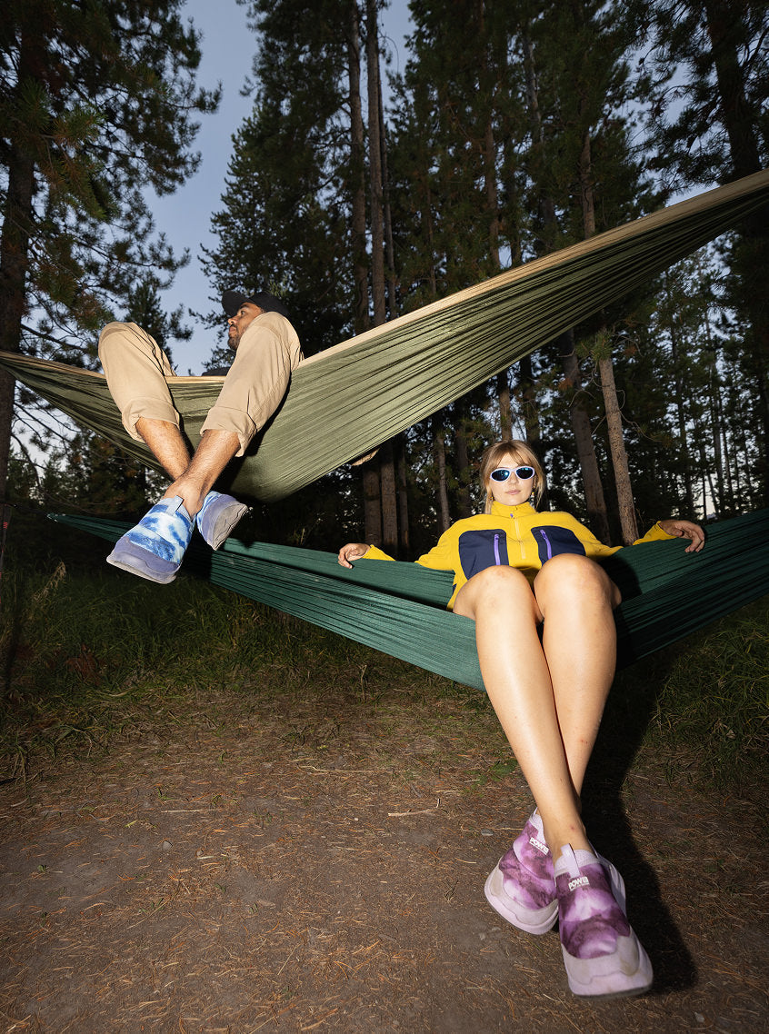 A man and a woman sit comfortably in a hammock, surrounded by nature, sharing a serene moment while both wearing bright colored slip-on shoes.