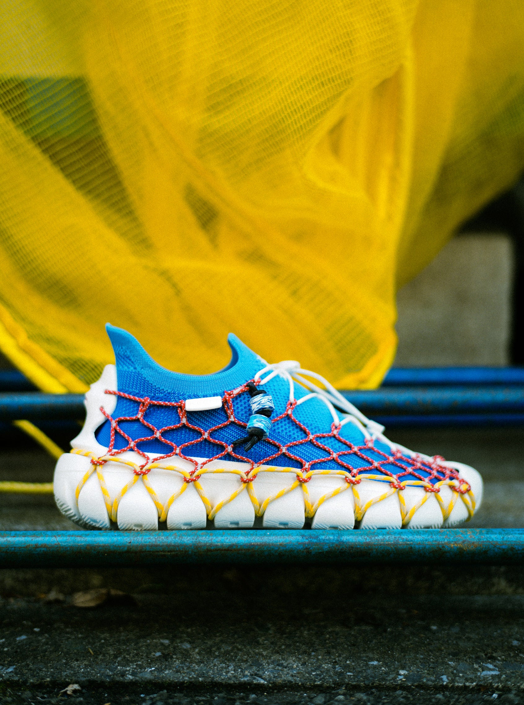 Product shot of the "Space Available" collaboration Uneek 360 Sneakers in blue and white, detailed with red and yellow lacing. 