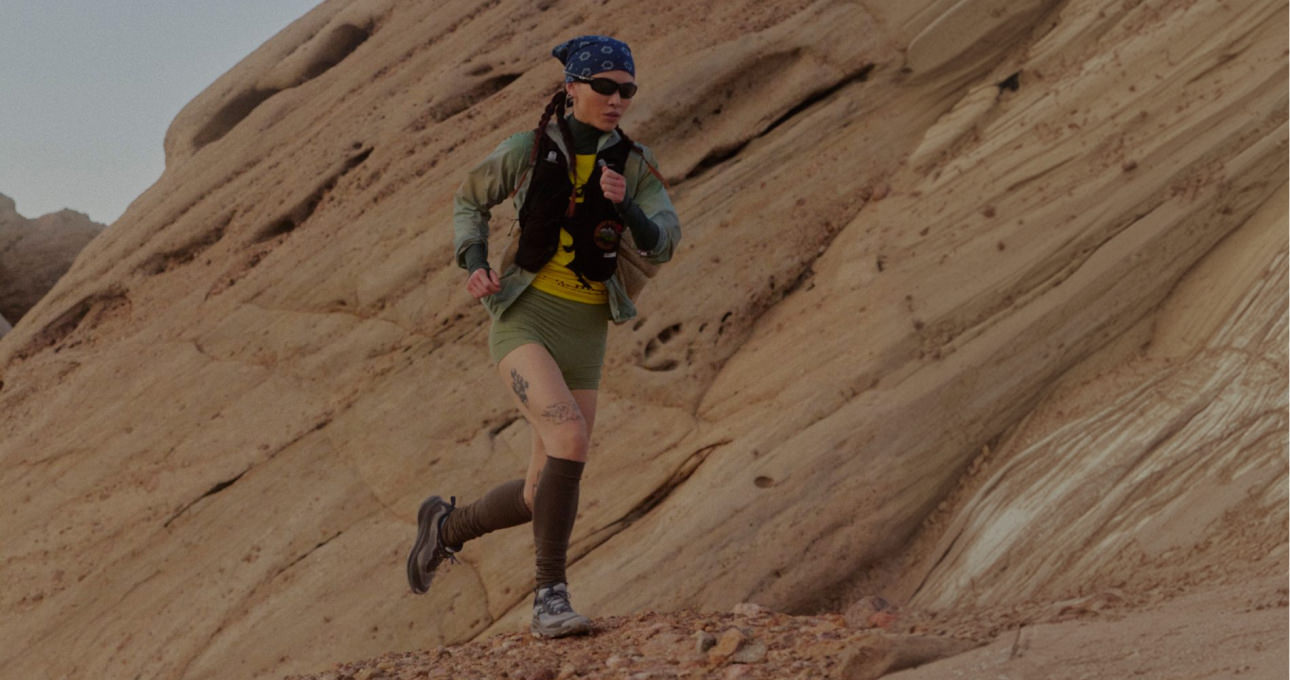 A woman wearing a bandana, running vest, and high socks in mid stride running along sandstone cliff. 