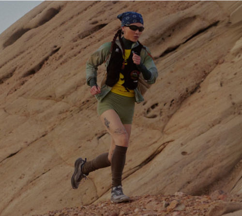 A woman wearing a bandana, running vest, and high socks in mid stride running along sandstone cliff. 