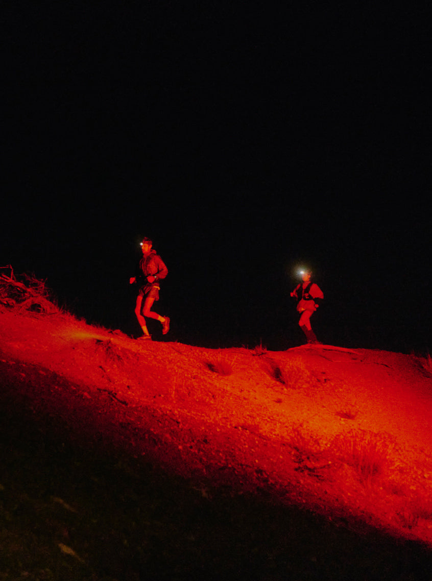Two trai runners in the dark making strides across a trail with their headlamps lighting the way in red light.
