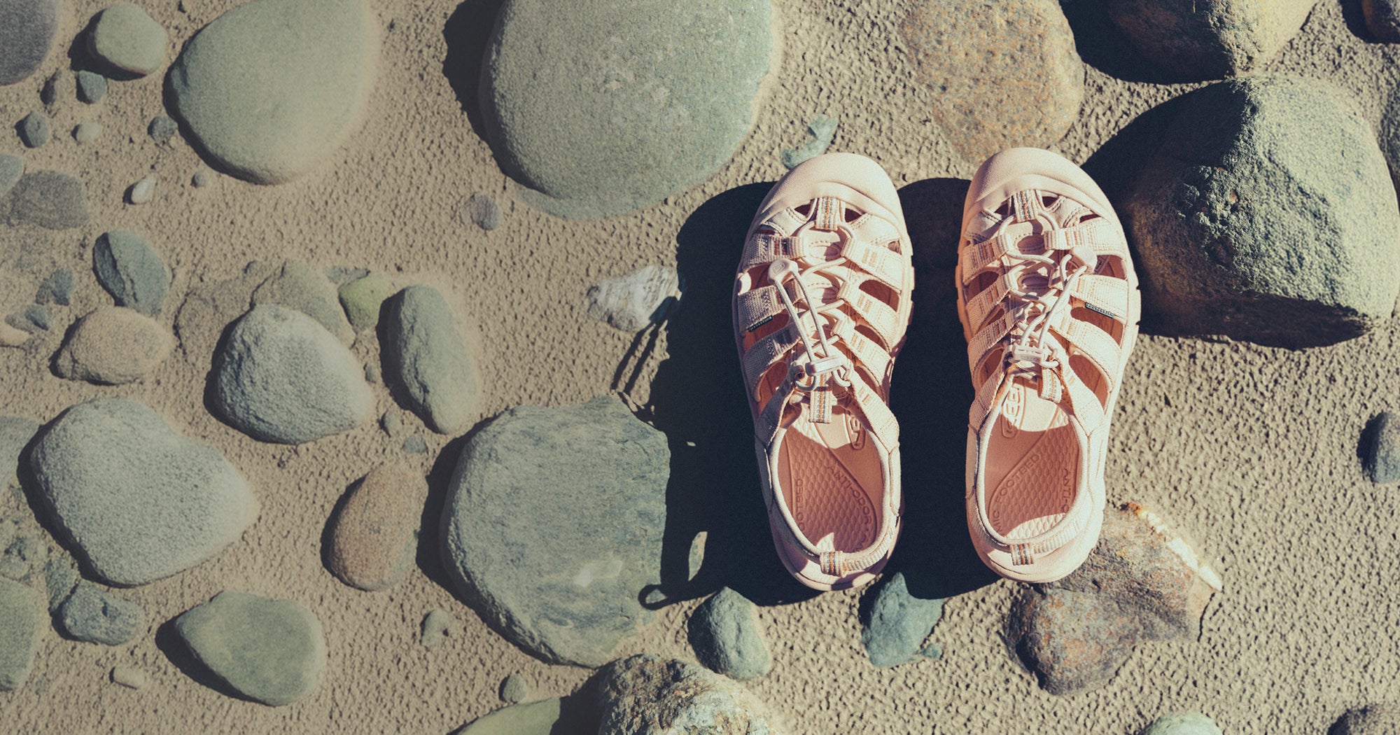A pair of monochrome rose colored women's newport sandals placed on a rocky and sandy beach. 