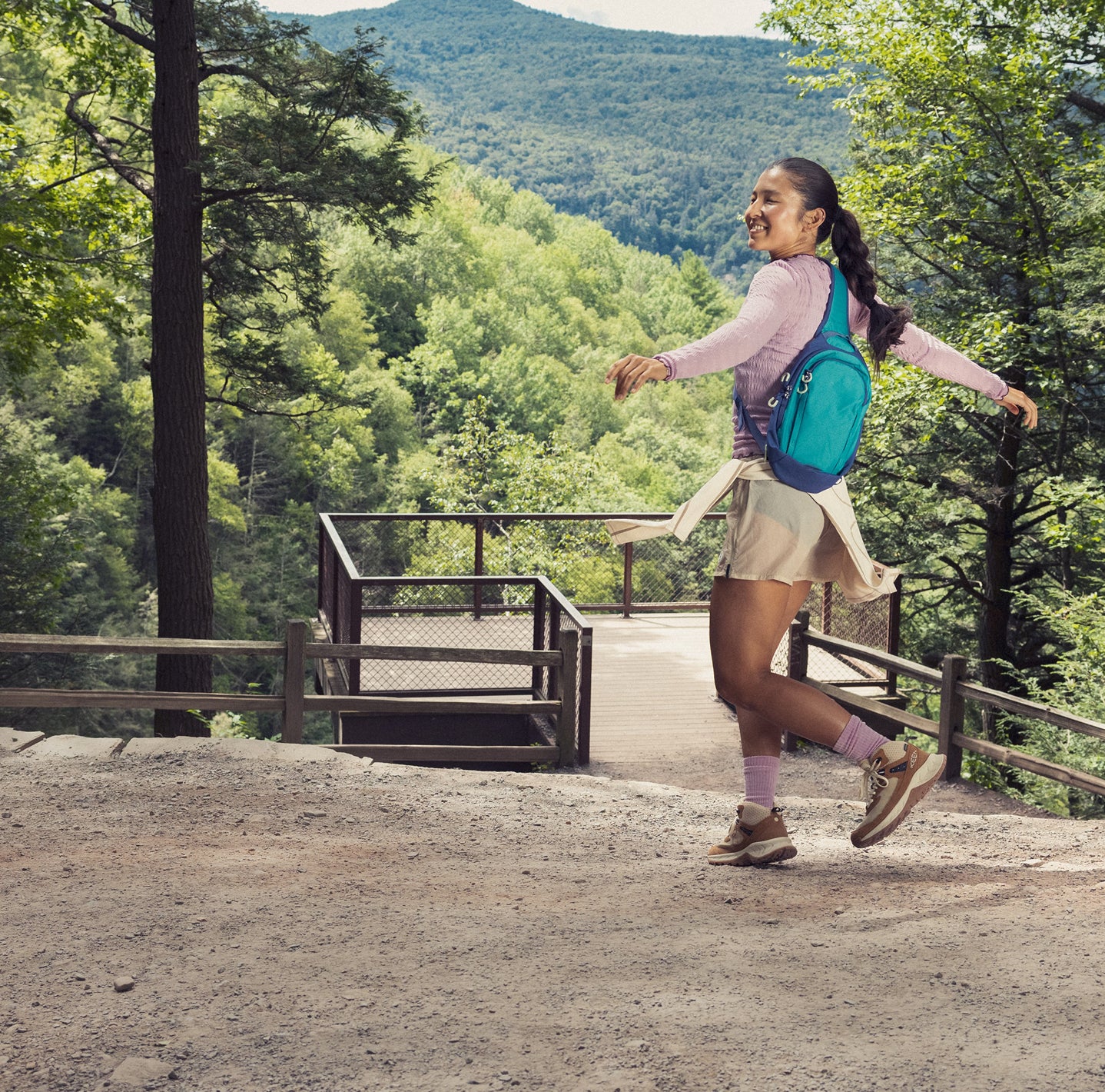 A woman in a cute purple hiking outfit with a teal backpack twirling in the trees while wearing brown and tan stylish HIghtrail hiking boots. 
