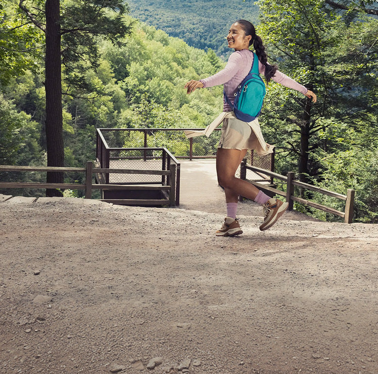 A woman in a cute purple hiking outfit with a teal backpack twirling in the trees while wearing brown and tan stylish HIghtrail hiking boots. 