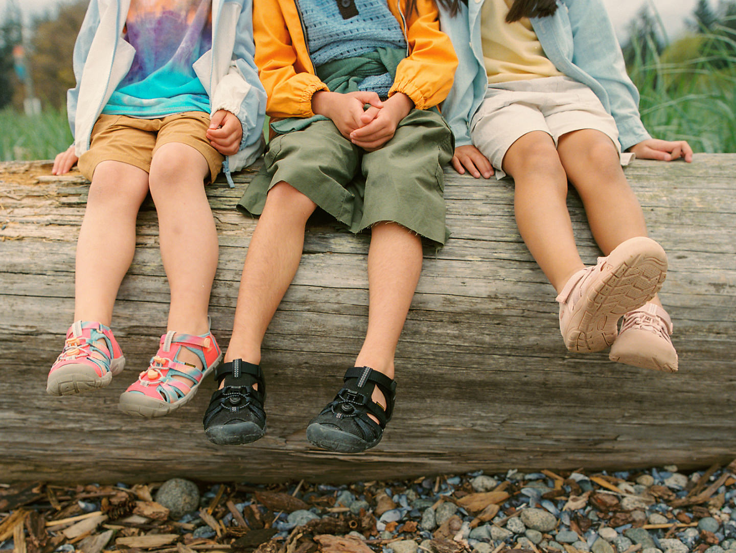 Three young kids sitting a log and dangling their feet off while wearing a variety of KEEN sandals with bungee laces and closed toe protection. 
