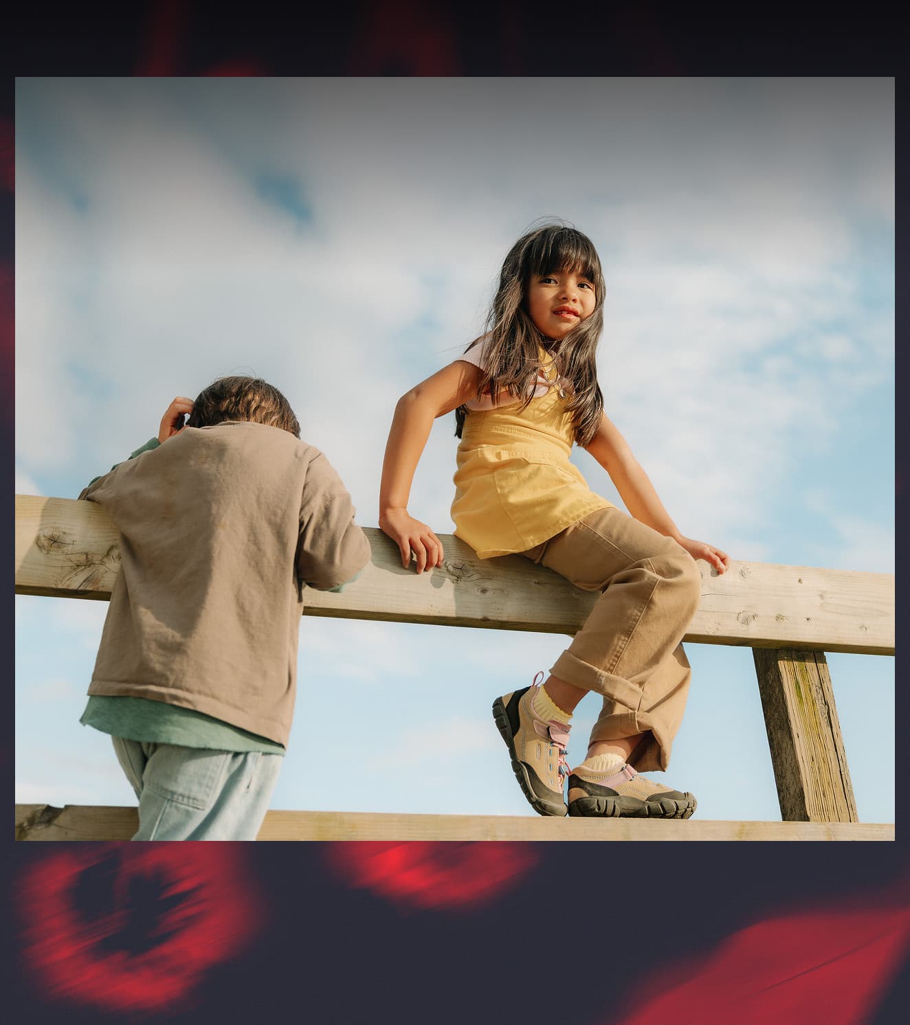 A young girl and boy sitting on a fence and wearing KEEN sneakers with hook-and-loop straps and bungee laces.