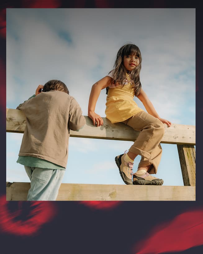 A young girl and boy sitting on a fence and wearing KEEN sneakers with hook-and-loop straps and bungee laces.