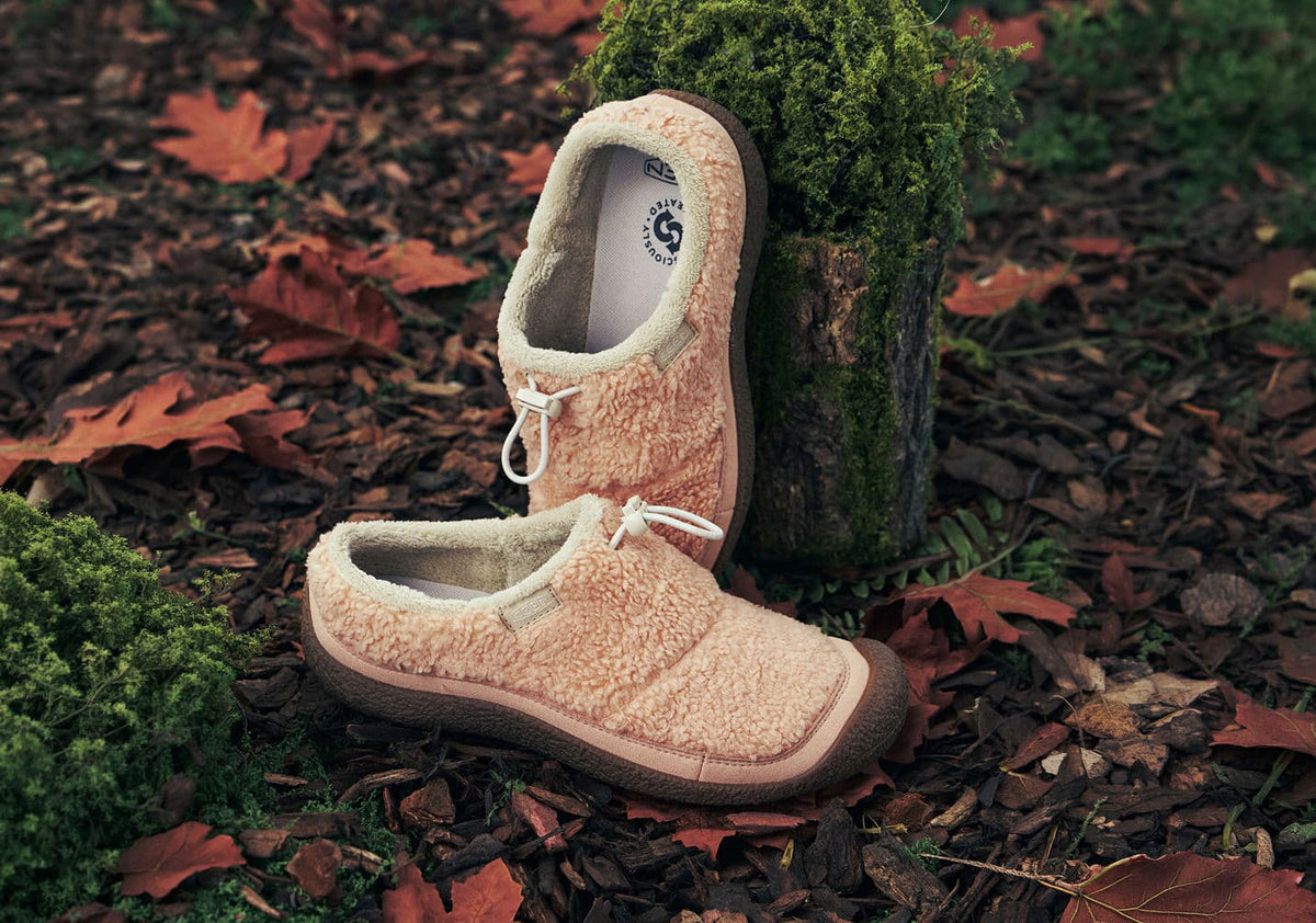 A pair of women's Howser slides with a pink, fleece upper and brown sole, perched up against a mossy tree.