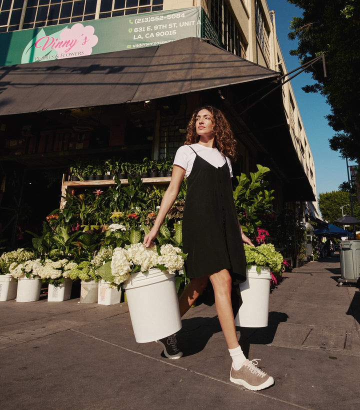 A woman in a black and white dress wearing KEEN MADE leather sneakers walking out of a floral shop with a pail of flowers in each hand.