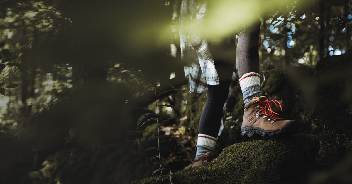 Person in leggings and flannel shirt hiking through moss covered forest in KEEN brown Pyrenees Boots with red laces.  