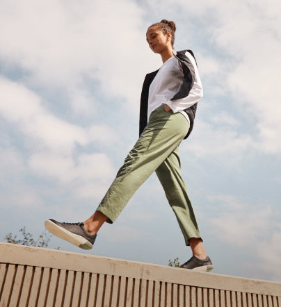 Woman wearing KNX Leather Sneakers and walking on top of wooden structure with clouds behind her. 