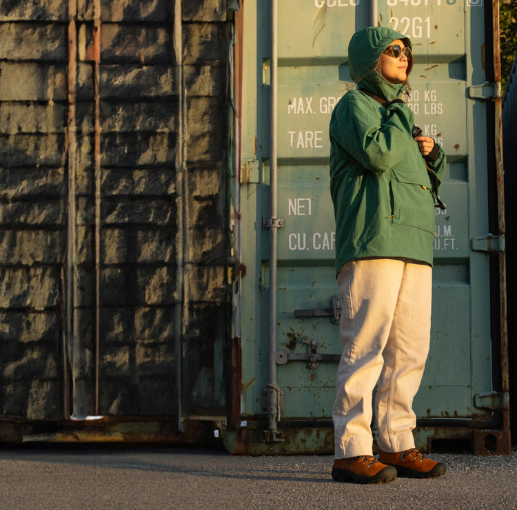 Person in green hooded sweatshirt wearing brown Pyrenees boots and stading in front of shipping container